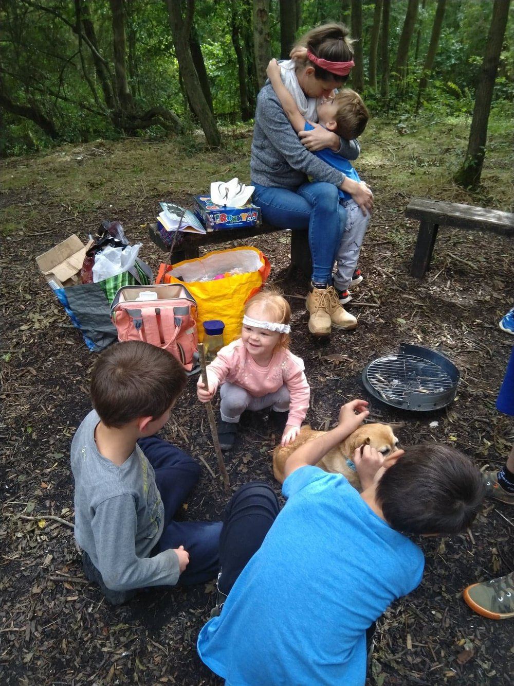 A parent embracing her child during a forest school session, with other children and a dog nearby